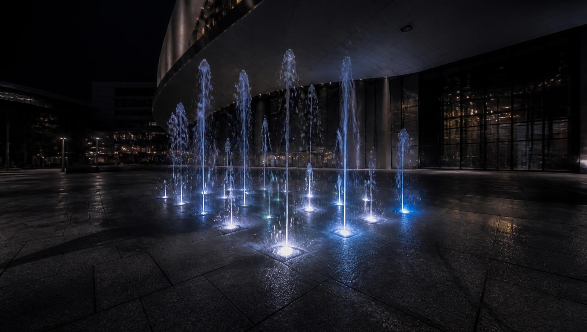 Illuminated dry deck fountain jets rising from a granite plaza at night